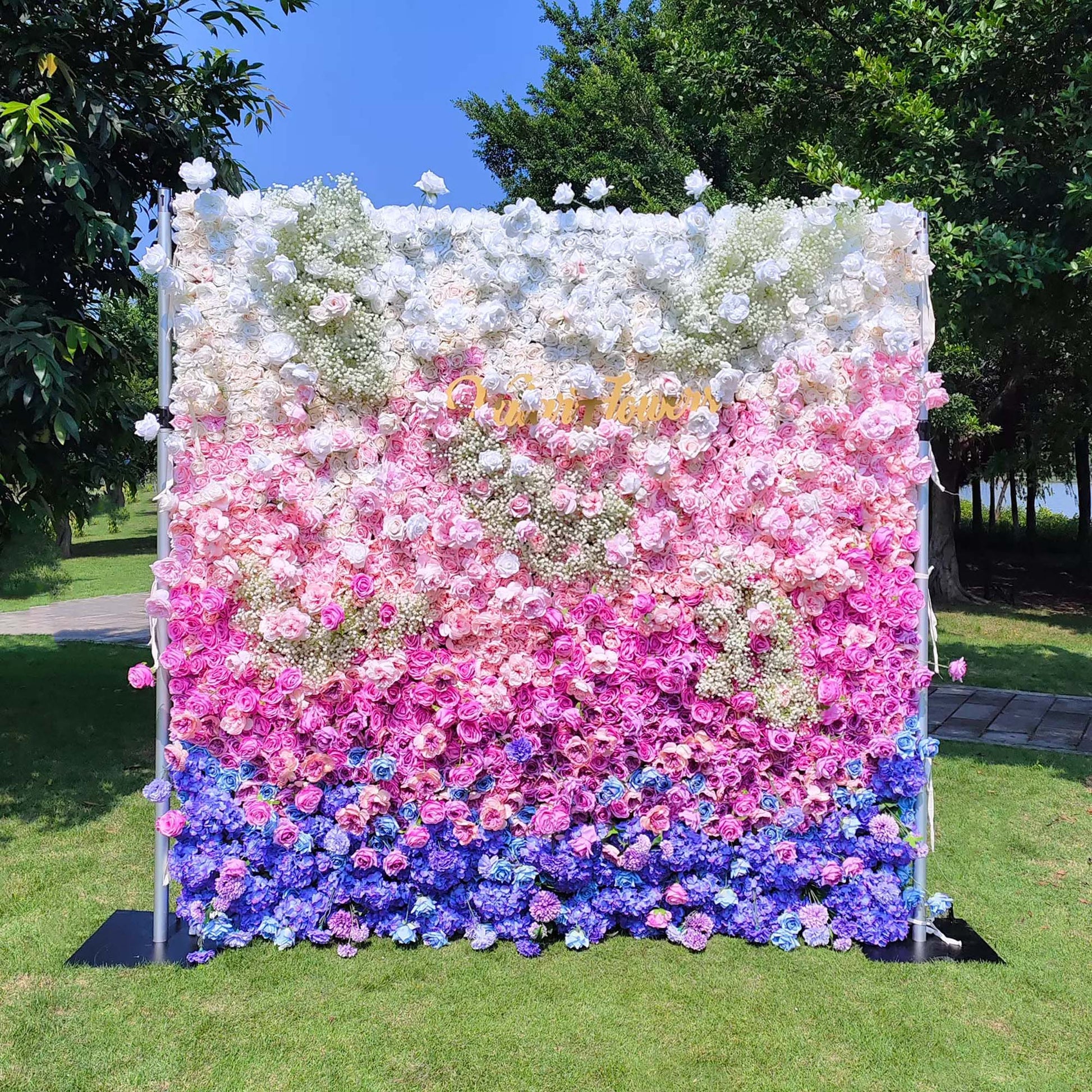 This photo features a lush floral wall with a gradient of white, pink, and blue. Composed of roses and hydrangeas, with baby's breath adding texture. Set outdoors on green grass with trees in the background, it has a dreamy, romantic style, ideal for weddings or photo - booth backdrops. 