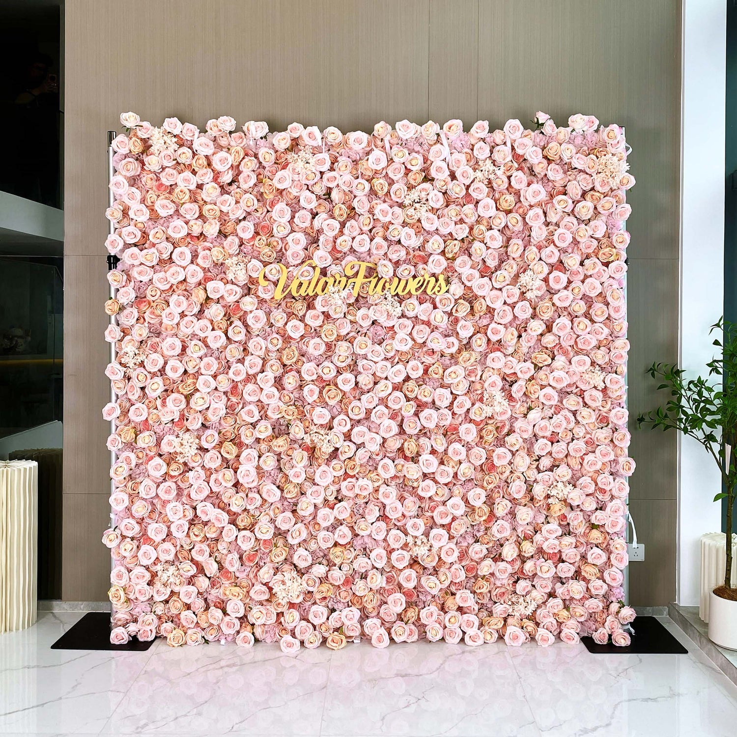 A large, square flower wall covered with an abundance of light pink roses. The text &quot;ValarFlowers&quot; is prominently displayed in the center, creating a romantic and elegant atmosphere.