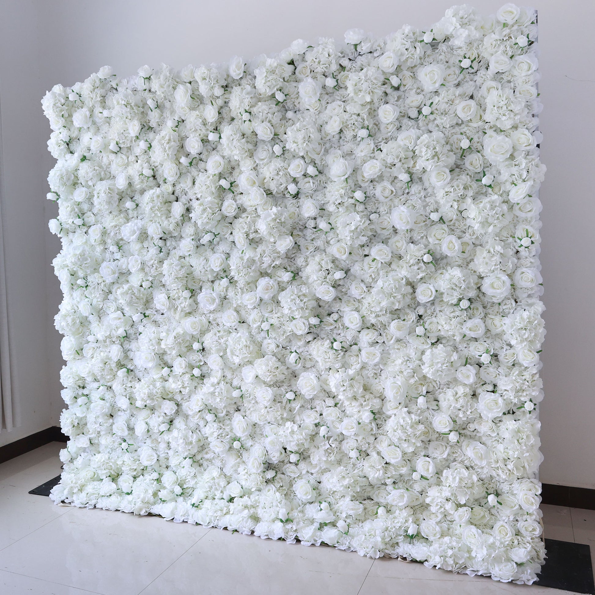 This image showcases a large, square floral wall indoors. It's densely covered with pure white flowers, creating a clean, elegant, and serene display. The flowers, including roses and other blooms, are tightly packed, giving a sense of opulence and purity. Set against a plain white wall with a shiny floor, the scene exudes an atmosphere of sophistication and natural beauty, perfect for decorative purposes in events or as a striking interior backdrop.