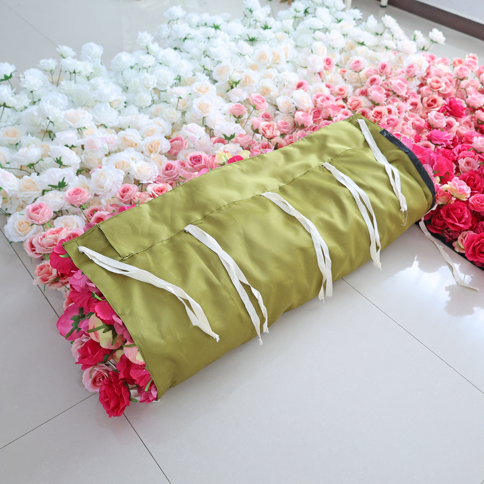 The image shows a rolled - up artificial flower wall with a green cover and white ties. The flower wall features a gradient of white, pink, and red roses, suggesting it's used for decorative purposes at events like weddings.