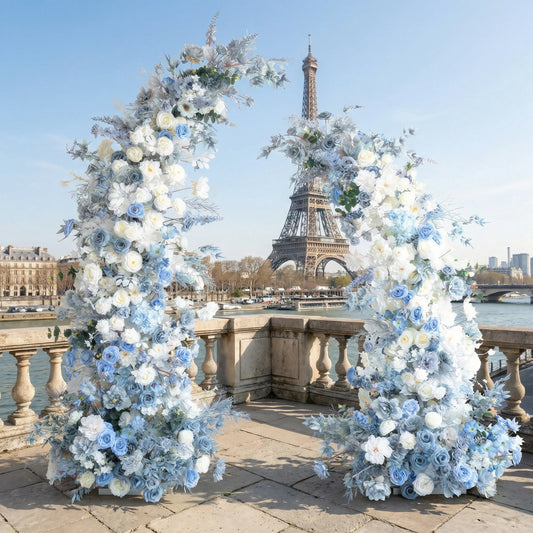 A dreamy, asymmetrical floral arch from **Valar Flowers**, crafted with a serene palette of sky-blue, white, and soft cream blooms—including roses, hydrangeas, and delicate foliage. Set on a Parisian riverside terrace with the iconic Eiffel Tower in the background, it creates a cool, elegant, and ethereal aesthetic ideal for luxury weddings and editorial photoshoots.