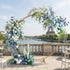 A sleek, gold circular floral arch from Valar Flowers, adorned with a serene palette of sky blue, navy, and creamy white blooms—including hydrangeas, roses, and delicate orchids—intertwined with cascading greenery. Set against the iconic Eiffel Tower and Seine River in Paris, it creates a sophisticated, romantic atmosphere perfect for a luxury wedding or special celebration.