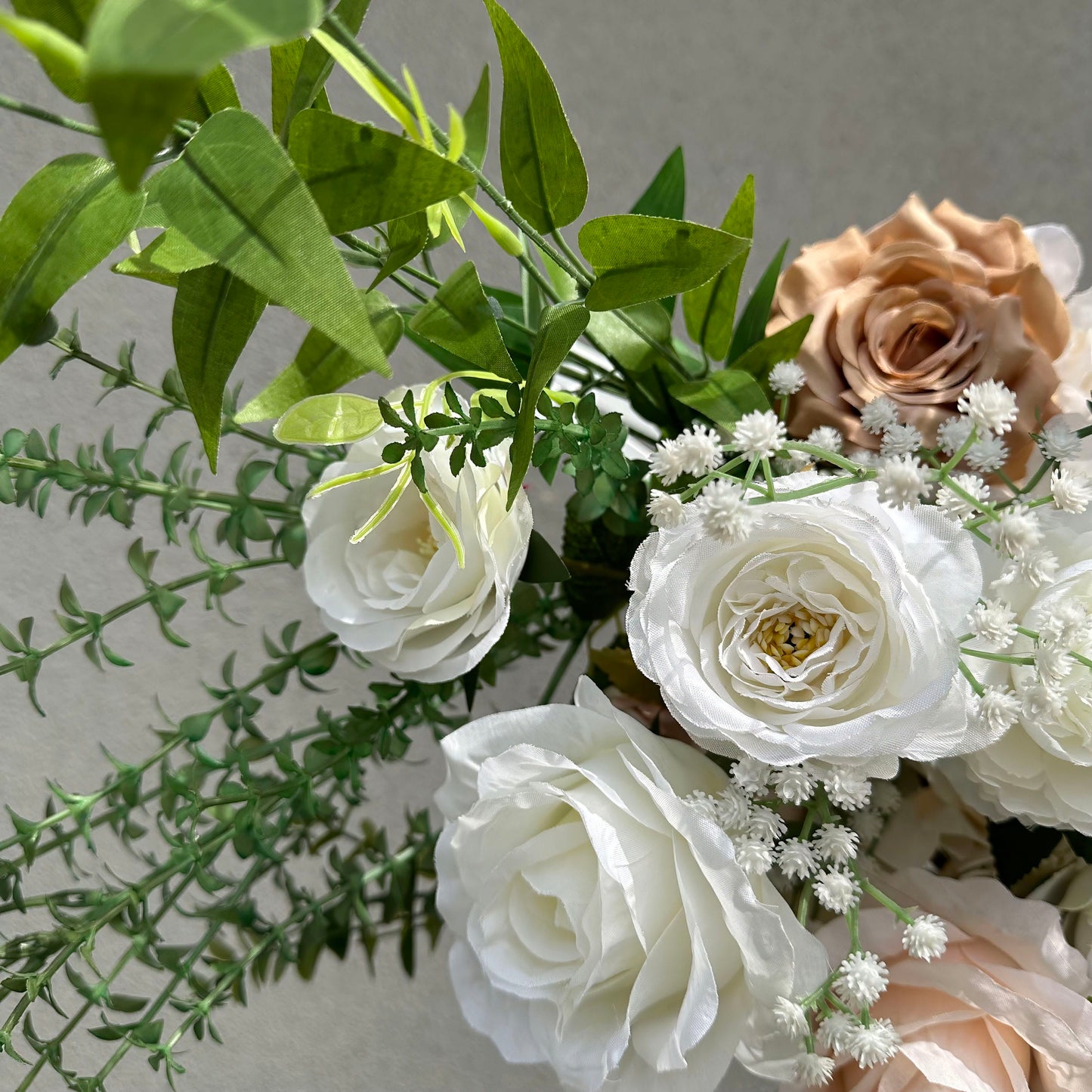 A close-up of a lush floral arrangement from Valar Flowers, featuring creamy white roses, delicate baby's breath, and a warm terracotta rose, all intertwined with vibrant green foliage. The soft, textured petals and fresh greenery create a romantic, elegant aesthetic, ideal for wedding decor, centerpieces, or special event accents.