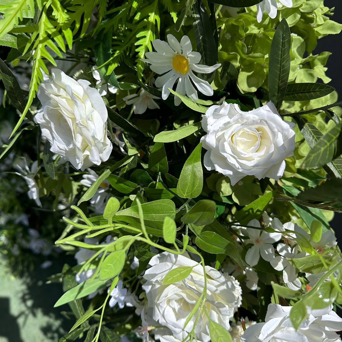 Close-up of a lush artificial floral arrangement from Valar Flowers, showcasing pristine white roses and cheerful daisies nestled in vibrant green foliage—including ferns and eucalyptus leaves. The sun-dappled texture highlights realistic petals and fresh-looking greenery, ideal for wedding backdrops, flower walls, or event decor accents.