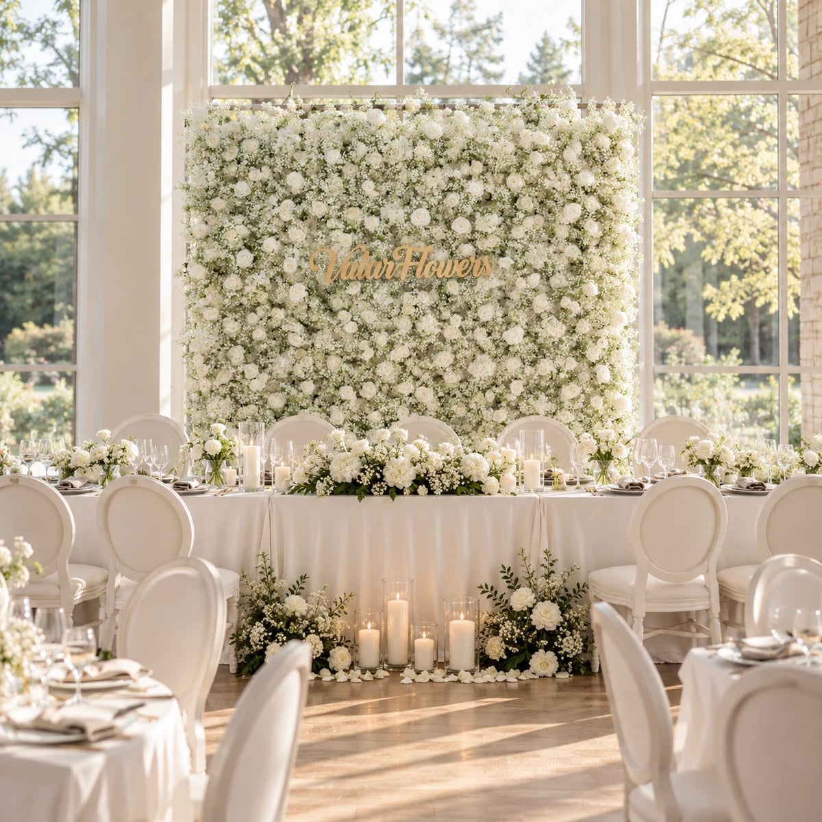 Luxury all-white baby’s breath and rose flower wall backdrop with elegant gold “ValarFlowers” signage for a wedding head table. Bright sunlit venue with floor-to-ceiling windows, ivory linen tables, white dining chairs, lush floral arrangements and glowing pillar candles. This timeless neutral floral setup works for wedding receptions, bridal events, brand photoshoots and classic minimalist wedding decor inspiration.