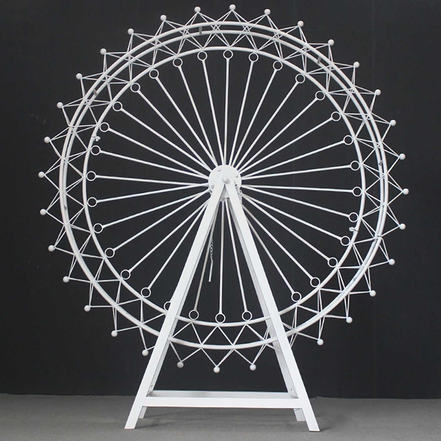 A large, white metal Ferris wheel decoration stands against a black backdrop. It has an intricate, geometric lattice design with circular spokes and small spheres. The triangular stand provides stability, showcasing a sleek, minimalist aesthetic—ideal for decorative event setups like weddings or parties. 