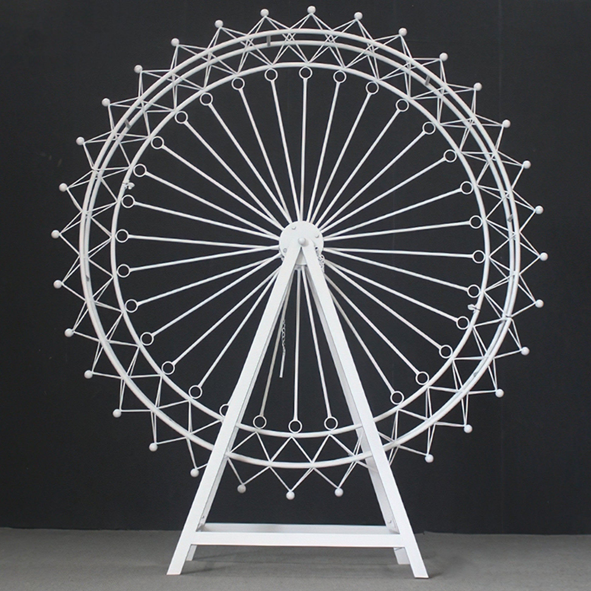 A large, white metal Ferris wheel decoration stands against a black backdrop. It has an intricate, geometric lattice design with circular spokes and small spheres. The triangular stand provides stability, showcasing a sleek, minimalist aesthetic—ideal for decorative event setups like weddings or parties. 
