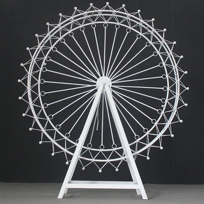 A large, white metal Ferris wheel decoration stands against a black backdrop. It has an intricate, geometric lattice design with circular spokes and small spheres. The triangular stand provides stability, showcasing a sleek, minimalist aesthetic—ideal for decorative event setups like weddings or parties. 