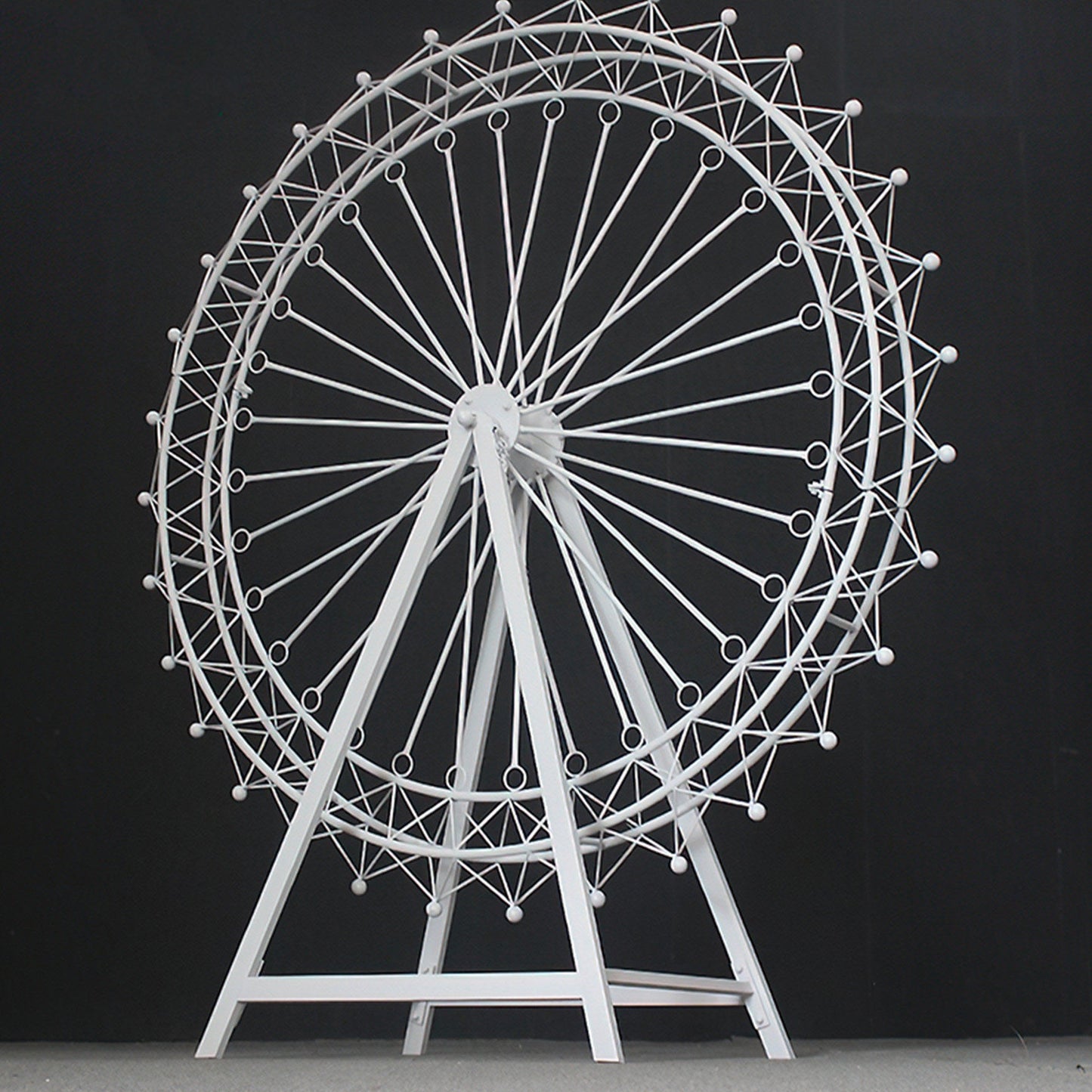 A large, white metal Ferris wheel decoration stands against a black backdrop. It features an intricate, geometric lattice design with circular spokes and small spheres. The triangular stand provides stability, showcasing a sleek, minimalist aesthetic, perfect for decorative event setups. 