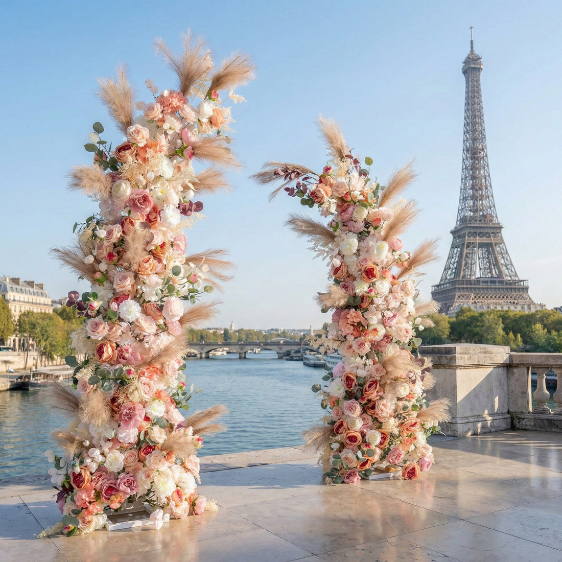 This is a romantic, asymmetrical floral arch from **Valar Flowers**, adorned with a soft palette of blush pink, peach, white blooms, and airy pampas grass, complemented by lush greenery. Set on a Parisian riverside terrace with the iconic Eiffel Tower in the background, it creates a dreamy, elegant, and picturesque setting ideal for wedding ceremonies, photo shoots, or special events.