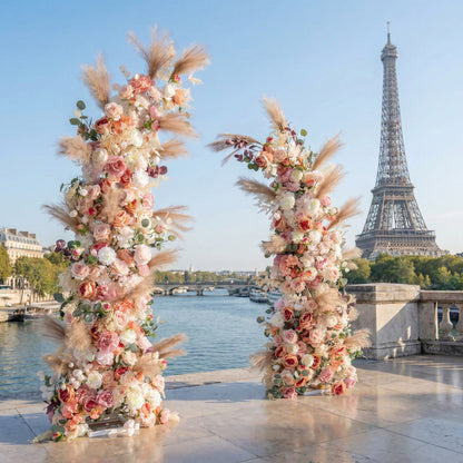 This is a romantic, asymmetrical floral arch from **Valar Flowers**, adorned with a soft palette of blush pink, peach, white blooms, and airy pampas grass, complemented by lush greenery. Set on a Parisian riverside terrace with the iconic Eiffel Tower in the background, it creates a dreamy, elegant, and picturesque setting ideal for wedding ceremonies, photo shoots, or special events.