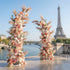 This is a romantic, asymmetrical floral arch from **Valar Flowers**, adorned with a soft palette of blush pink, peach, white blooms, and airy pampas grass, complemented by lush greenery. Set on a Parisian riverside terrace with the iconic Eiffel Tower in the background, it creates a dreamy, elegant, and picturesque setting ideal for wedding ceremonies, photo shoots, or special events.