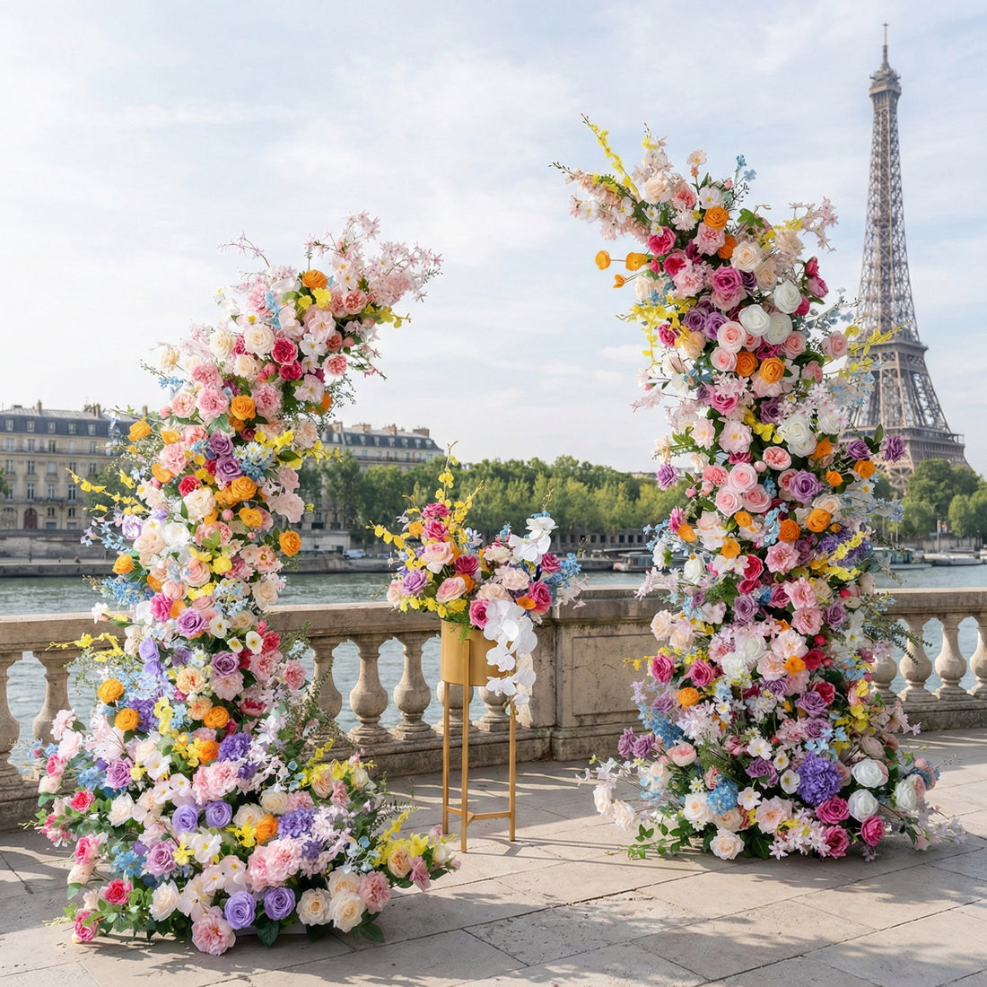 A vibrant, asymmetrical floral arch from **Valar Flowers**, bursting with a rainbow of blooms—from soft pink and peach to bold red, sunny yellow, and deep purple—intertwined with lush greenery. Set on a Parisian riverside terrace with the iconic Eiffel Tower in the background, it creates a joyful, romantic, and picturesque aesthetic ideal for luxury weddings and editorial photoshoots.