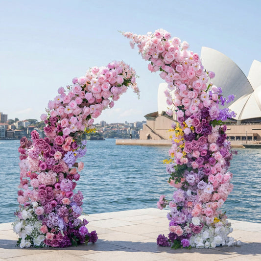 This romantic floral arch from Valar Flowers is a lush tapestry of blush pink, lavender, and white blooms—including roses, hydrangeas, and delicate wisteria—arranged in two cascading columns. Set against the iconic Sydney Opera House and the glistening harbor, it evokes a fresh, romantic atmosphere ideal for a coastal wedding, engagement photoshoot, or elegant celebration.