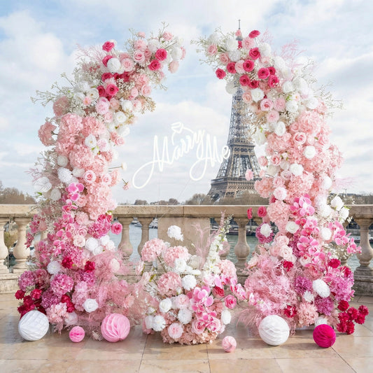 This romantic heart-shaped floral arch from **Valar Flowers** is a lush tapestry of blush pink, deep rose, and creamy white blooms—including roses, peonies, and delicate filler flowers. Suspended at its center is a glowing "Marry Me" neon sign. Set against the iconic Eiffel Tower in Paris, the design evokes a dreamy, romantic atmosphere ideal for a heartfelt proposal or engagement photoshoot.