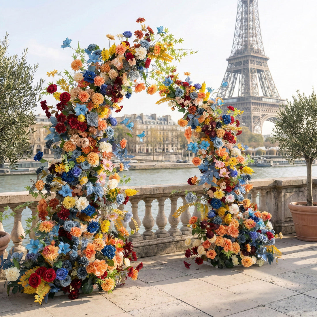 This is a vibrant, asymmetrical floral arch from Valar Flowers, bursting with a rich mix of blooms in blue, yellow, orange, red, pink, and white, complemented by lush greenery. Set on a Parisian riverside terrace with the iconic Eiffel Tower in the background, it creates a bold, cheerful, and eye-catching aesthetic ideal for weddings, photo shoots, or special events.