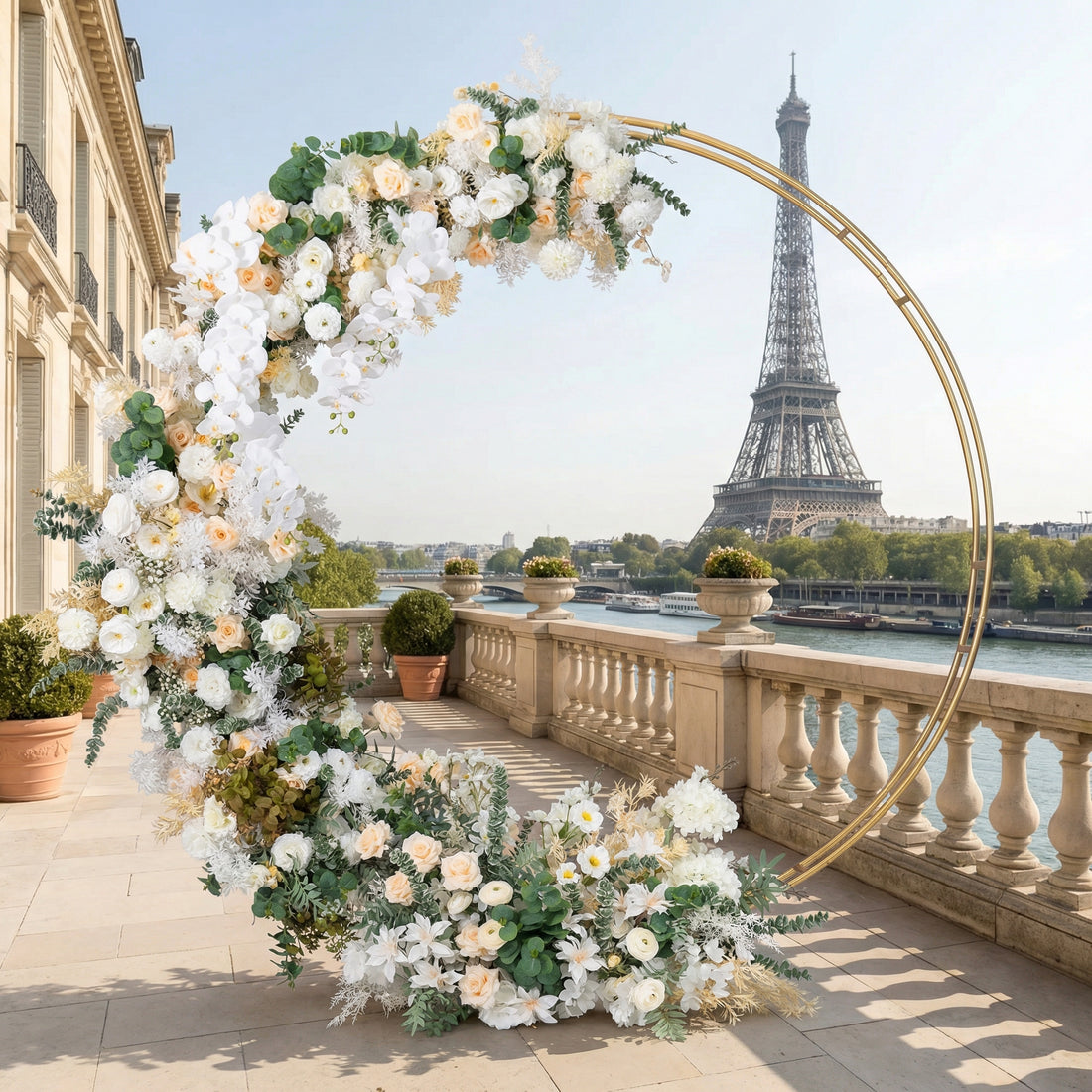 A sleek, gold circular floral arch from Valar Flowers, adorned with a soft palette of creamy white, blush pink, and warm peach blooms—including roses, peonies, and delicate orchids—intertwined with lush greenery. Set against the iconic Eiffel Tower and Seine River in Paris, it creates an elegant, romantic atmosphere perfect for a luxury wedding or special celebration.