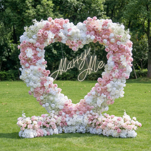 This romantic outdoor heart-shaped floral arch bursts with a dreamy blend of blush pink, deep rose, and crisp white blooms, including roses, hydrangeas, and delicate baby's breath. At its center, a glowing "Marry Me" neon sign takes the spotlight. Set on a lush green lawn framed by leafy trees, the arrangement exudes timeless romance, making it the perfect focal point for a heartfelt proposal, engagement photoshoot, or romantic celebration.