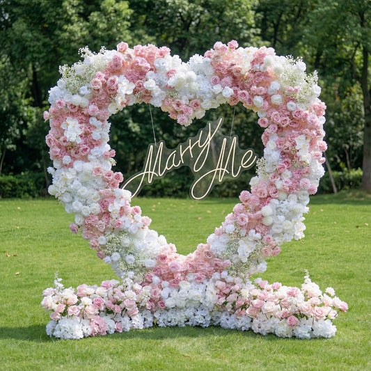 This romantic outdoor heart-shaped floral arch bursts with a dreamy blend of blush pink, deep rose, and crisp white blooms, including roses, hydrangeas, and delicate baby's breath. At its center, a glowing "Marry Me" neon sign takes the spotlight. Set on a lush green lawn framed by leafy trees, the arrangement exudes timeless romance, making it the perfect focal point for a heartfelt proposal, engagement photoshoot, or romantic celebration.
