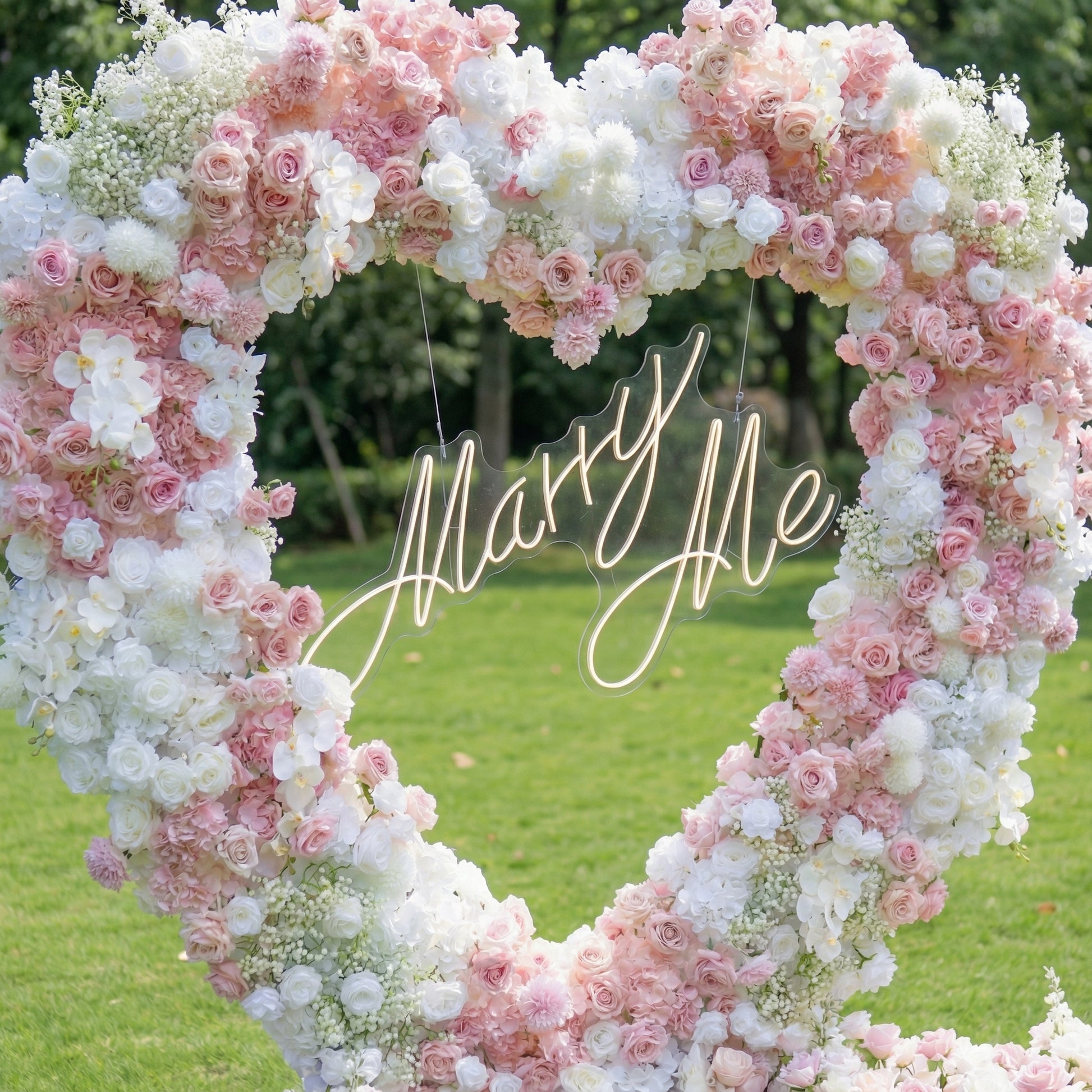 This close-up captures a romantic heart-shaped floral arch, crafted from a lush blend of blush pink, deep rose, and crisp white blooms—including roses, hydrangeas, orchids, and delicate baby's breath. At its center, a glowing "Marry Me" neon sign takes the spotlight. The dense, layered design creates a dreamy, three-dimensional texture, evoking timeless romance ideal for a heartfelt proposal or engagement photoshoot.