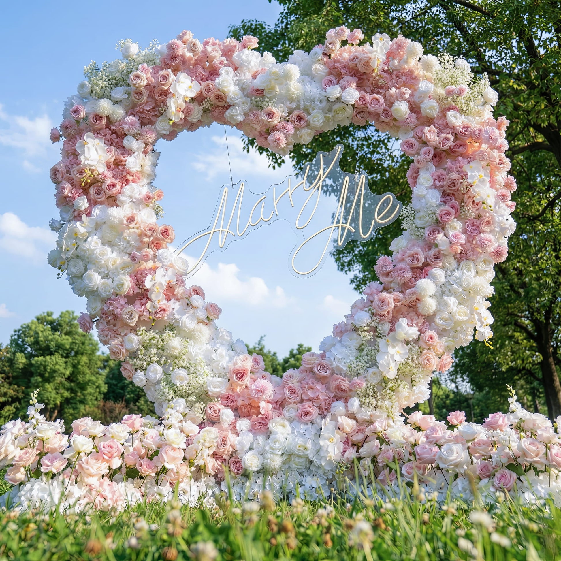This romantic heart-shaped floral arch, set against a bright blue sky and lush green trees, is crafted from a lush blend of blush pink, deep rose, and crisp white blooms—including roses, hydrangeas, orchids, and delicate baby's breath. At its center, a glowing "Marry Me" neon sign takes the spotlight. The dense, layered design creates a dreamy, three-dimensional texture, evoking timeless romance ideal for a heartfelt proposal or engagement photoshoot.