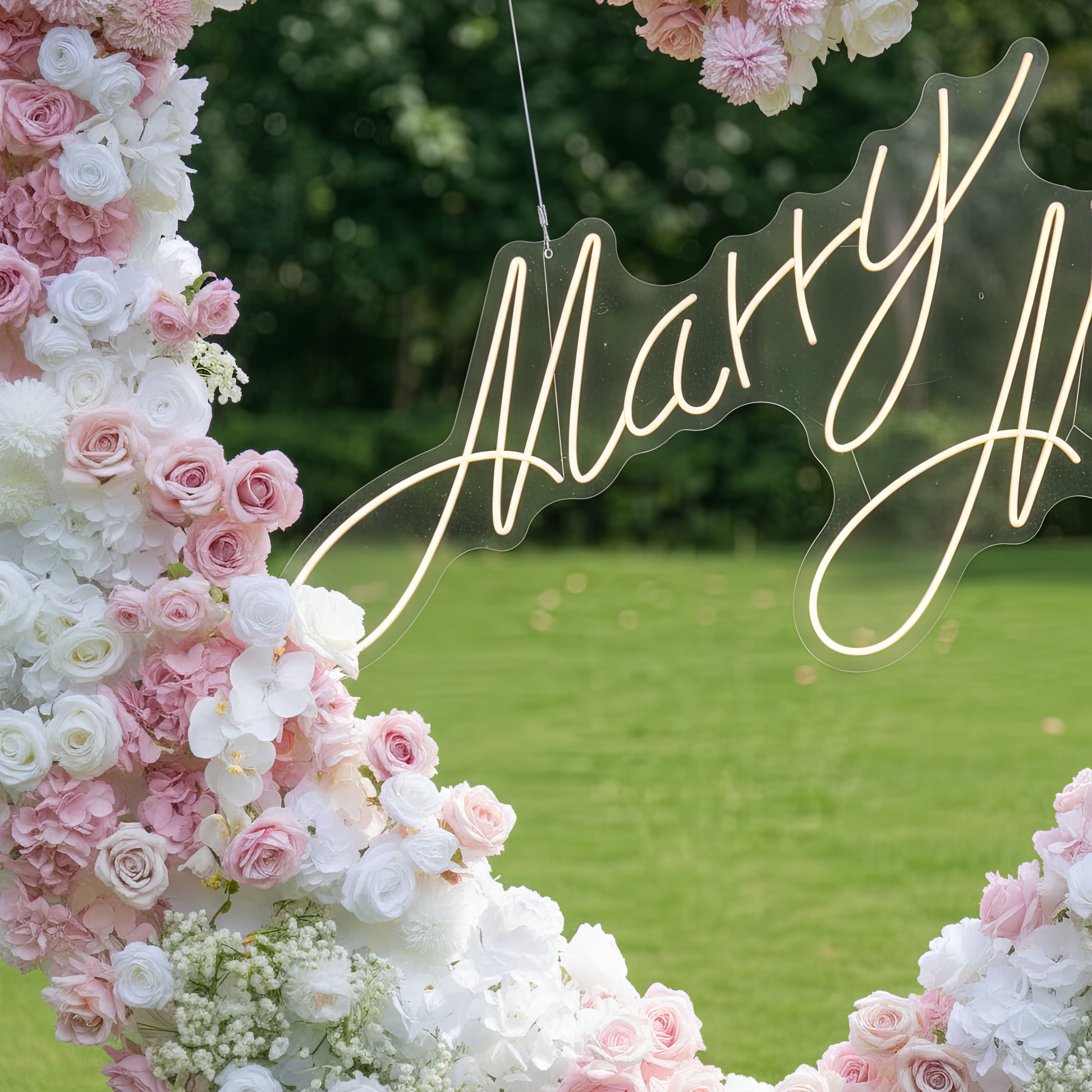 This close-up captures a romantic heart-shaped floral arch, crafted from a lush blend of blush pink, deep rose, and crisp white blooms—including roses, hydrangeas, orchids, and delicate baby's breath. A glowing "Marry Me" neon sign hangs at its center, suspended by clear wire. The dense, layered design creates a dreamy, three-dimensional texture, evoking timeless romance ideal for a heartfelt proposal or engagement photoshoot. Set against a lush green lawn and leafy trees, it exudes a soft, romantic atmosph