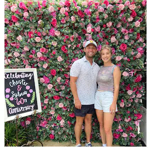 This photo captures a cheerful couple posing in front of a vibrant, lush flower wall backdrop. The background is completely covered in dense artificial roses in varying shades of pink and deep red, interspersed with green leaves, creating a romantic, full wall of blooms.
