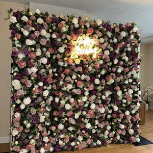 This stunning flower wall by Valar Flowers is a lush, vibrant backdrop featuring dense artificial blooms in soft pink, crisp white, rich purple, and peach hues, complemented by green foliage. A glowing neon sign adds a warm, celebratory touch, making it perfect for events like weddings, anniversaries, or parties. The wall’s full, layered design creates an elegant, romantic focal point, ideal for photo opportunities and special occasions.