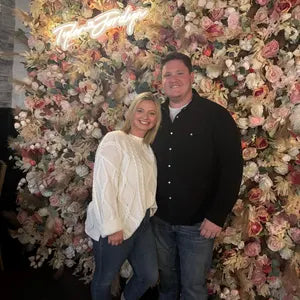 Two people posing in front of a lush floral wall with 'The Joinery' sign.