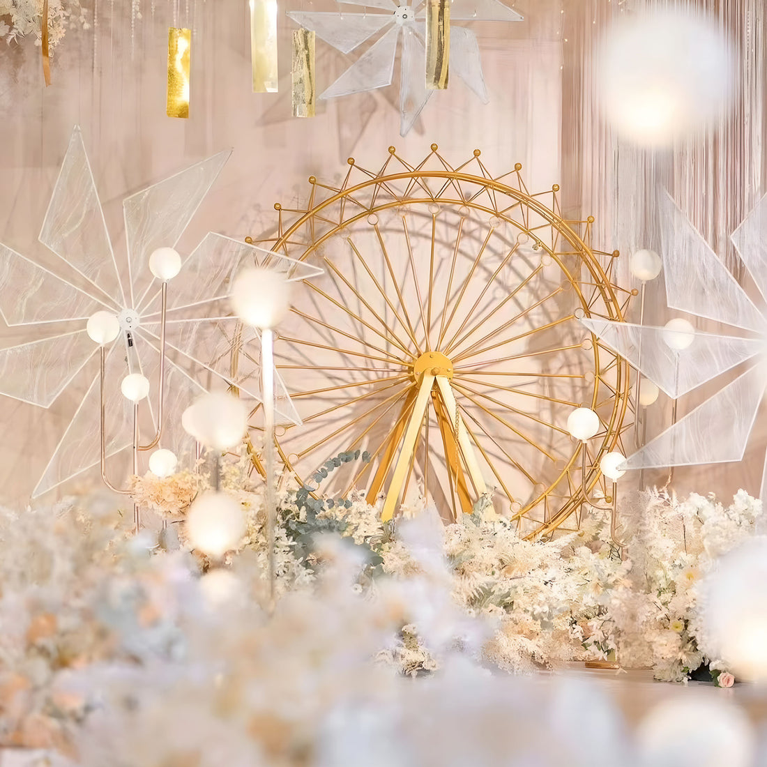 A dreamy event setup features a large, golden Ferris wheel as the centerpiece. Surrounding it are white, geometric decor pieces, glowing orbs, and lush floral arrangements in soft tones. Hanging gold and translucent elements add elegance, creating a romantic, whimsical atmosphere. 