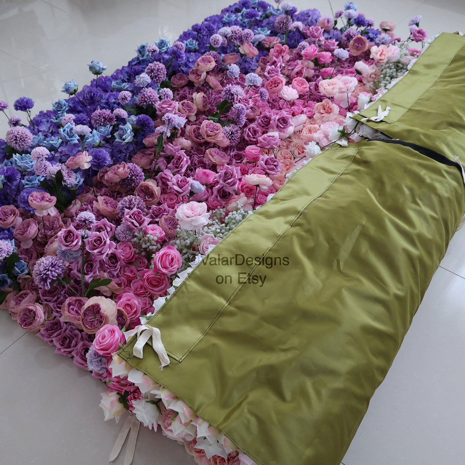 This photo displays a large floral arrangement on a tiled floor. It features a gradient of purple, pink, and white flowers, including roses, hydrangeas, and pom - pom flowers. Part is covered by an olive - green fabric. Ideal for weddings or events, it has a romantic style, with &quot;ValarDesigns on Etsy&quot; text visible. 