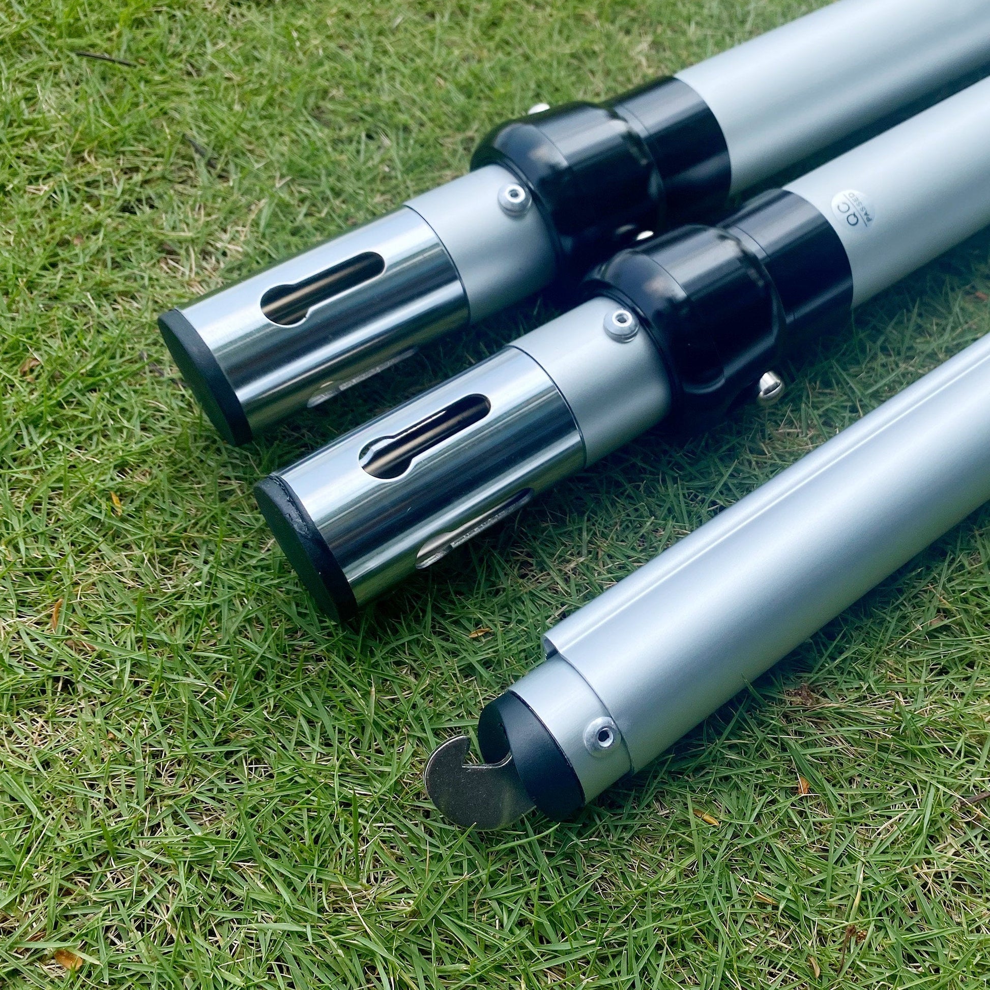Three silver metal poles with black connectors and end - caps rest on green grass. The poles have visible adjustment holes and a hooked end on one of them, suggesting they're parts of a modular setup. 