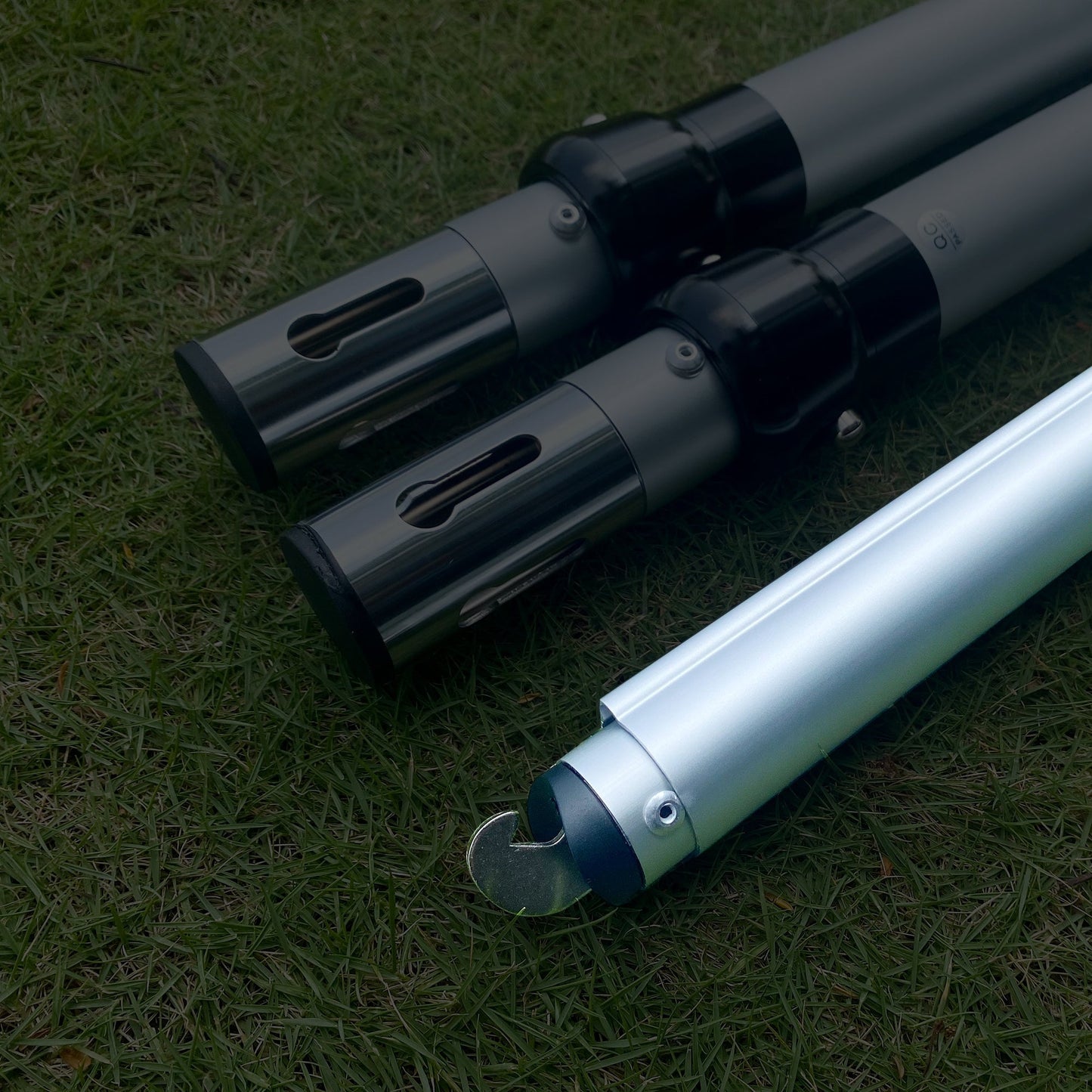 The image displays three metal poles on a grassy background. Two poles have black connectors with rectangular cut - outs, while the third has a hooked end, indicating they could be parts of an adjustable setup. 