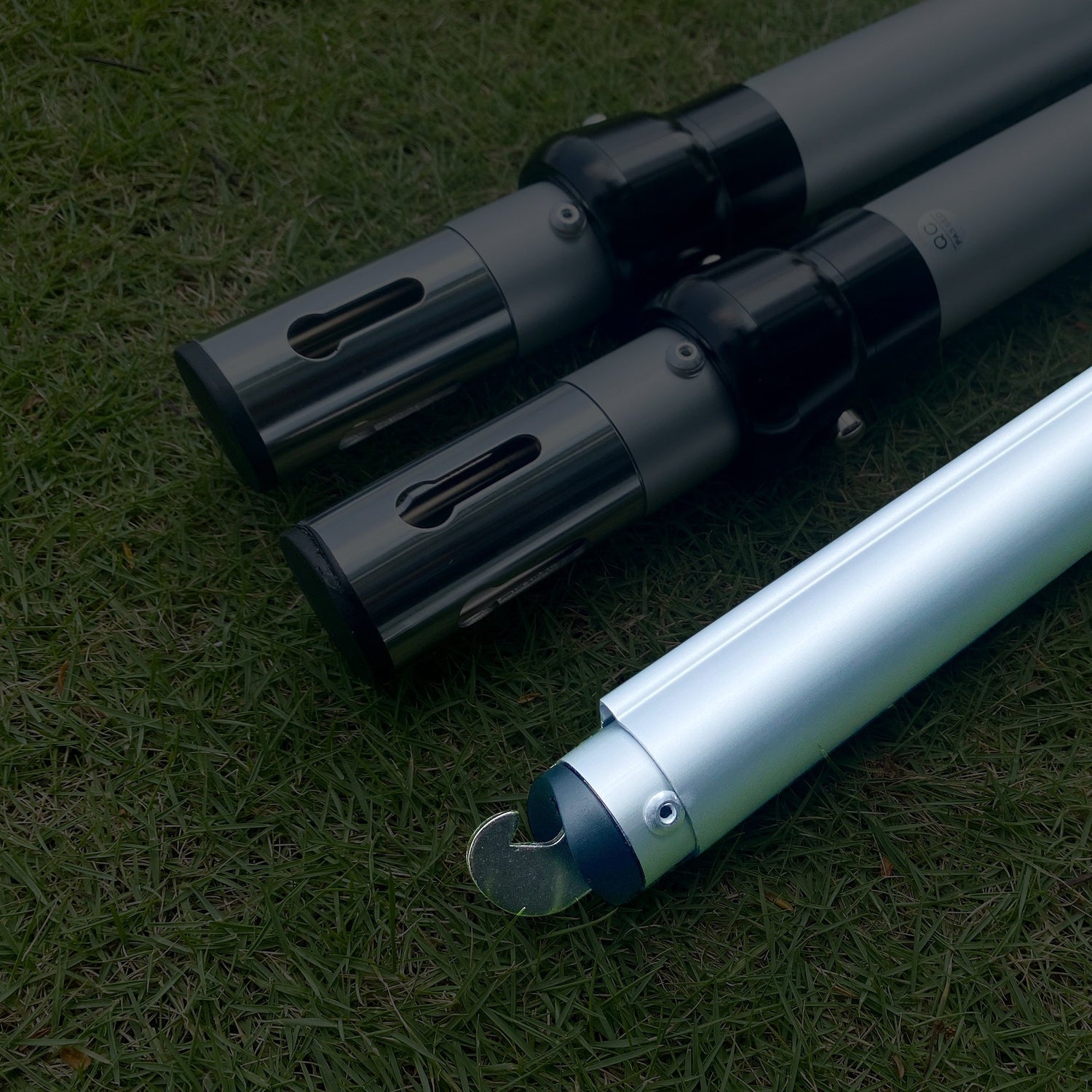 The image displays three metal poles on a grassy background. Two poles have black connectors with rectangular cut - outs, while the third has a hooked end, indicating they could be parts of an adjustable setup. 