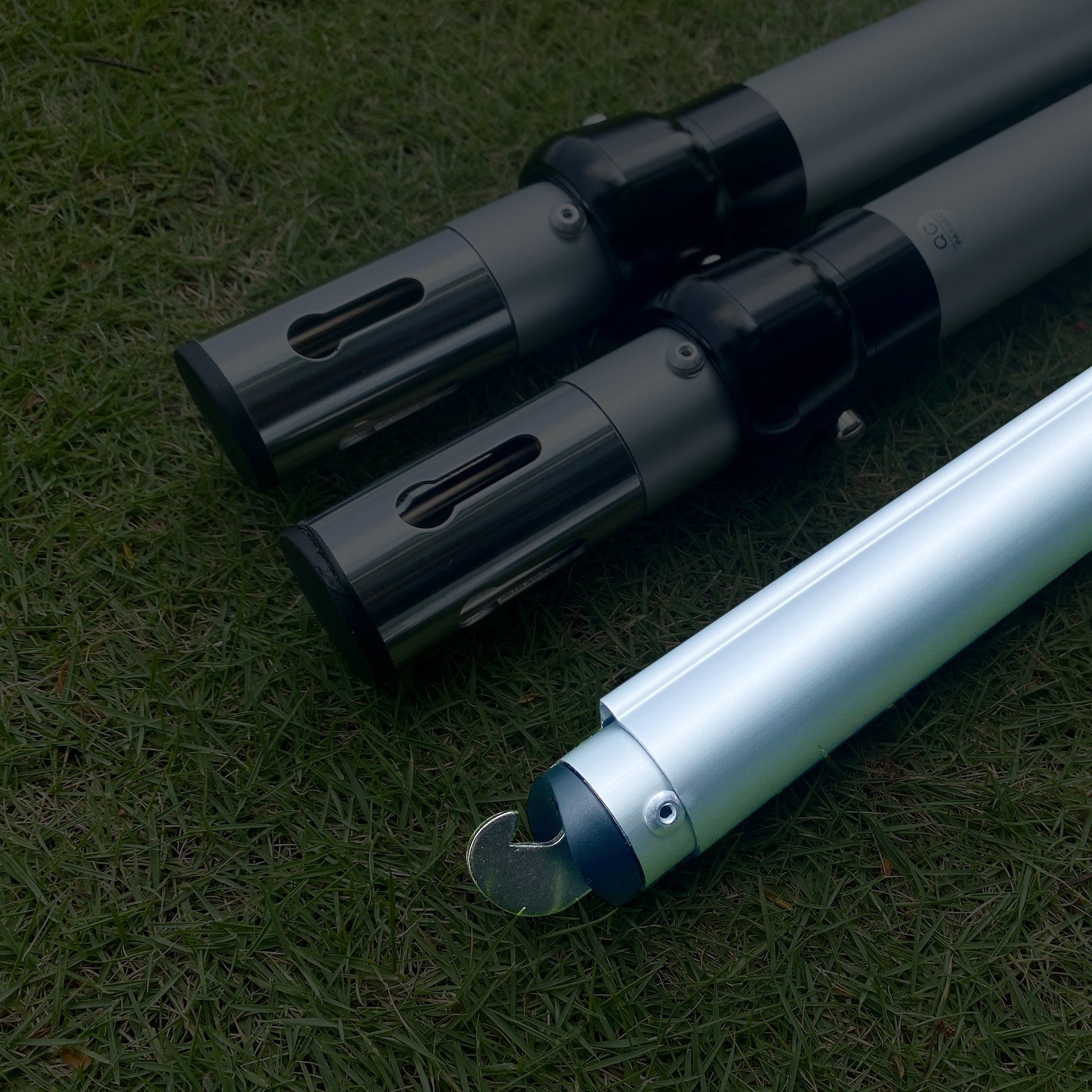 The image displays three metal poles on a grassy background. Two poles have black connectors with rectangular cut - outs, while the third has a hooked end, indicating they could be parts of an adjustable setup. 