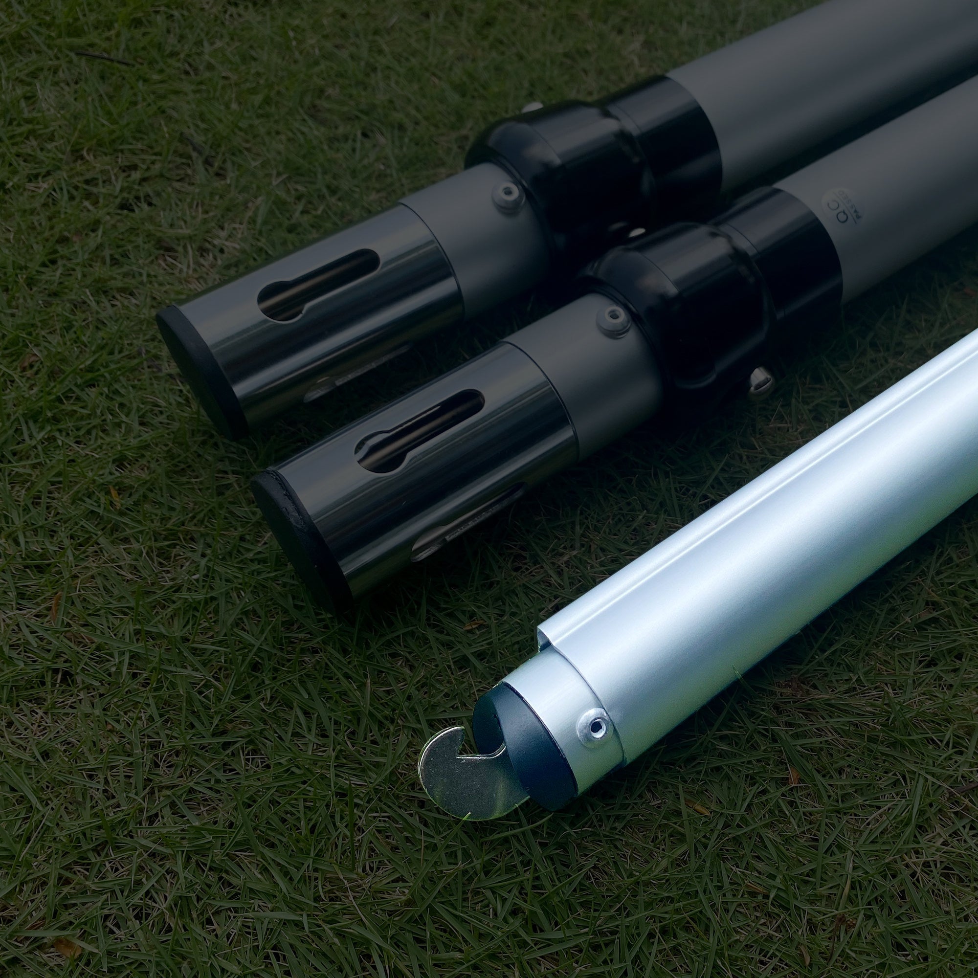 The image displays three metal poles on a grassy background. Two poles have black connectors with rectangular cut - outs, while the third has a hooked end, indicating they could be parts of an adjustable setup. 