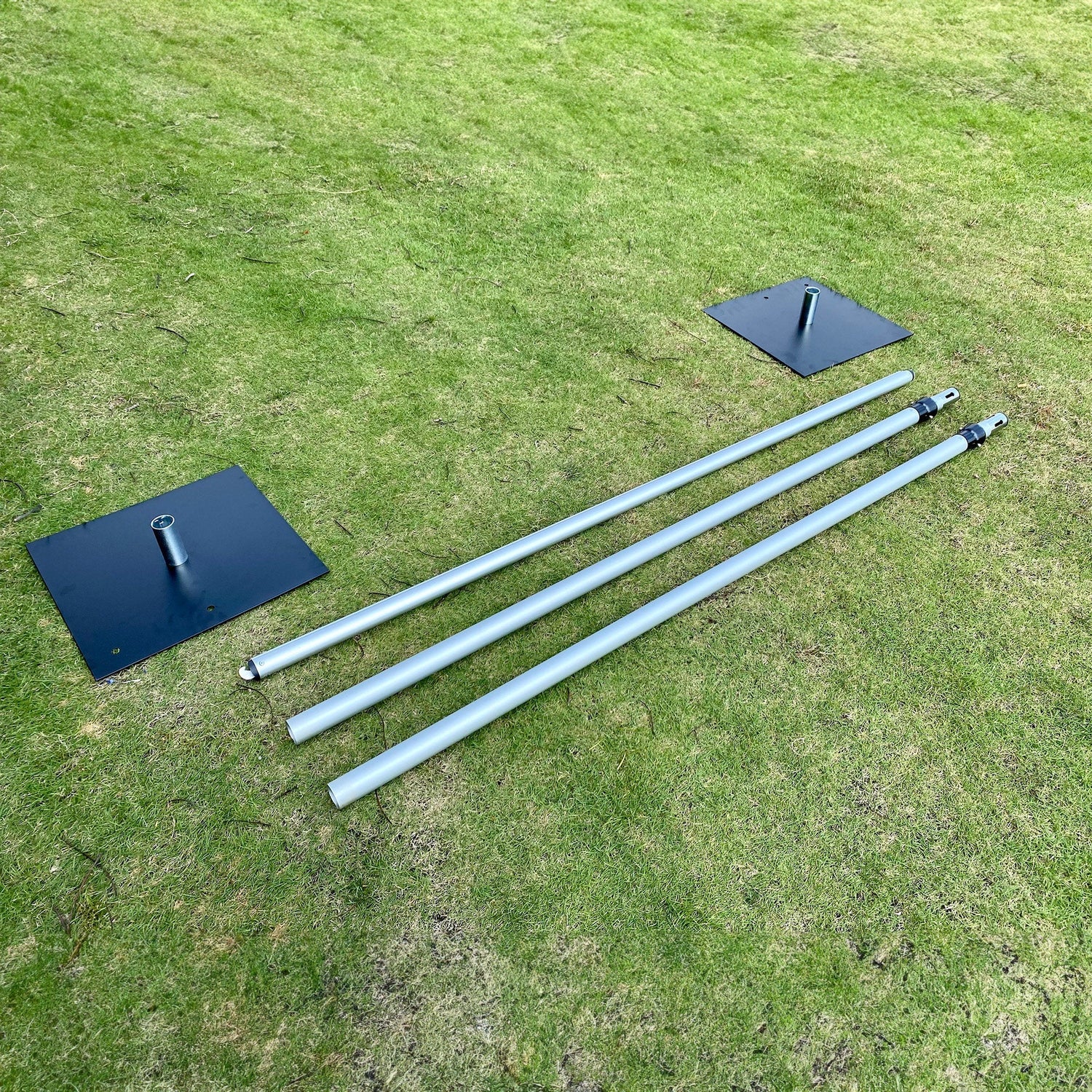 The image shows parts of a metal structure on a grassy surface. There are two black square bases with vertical metal sockets and three silver poles, some with black connectors, likely for assembling a larger framework. 
