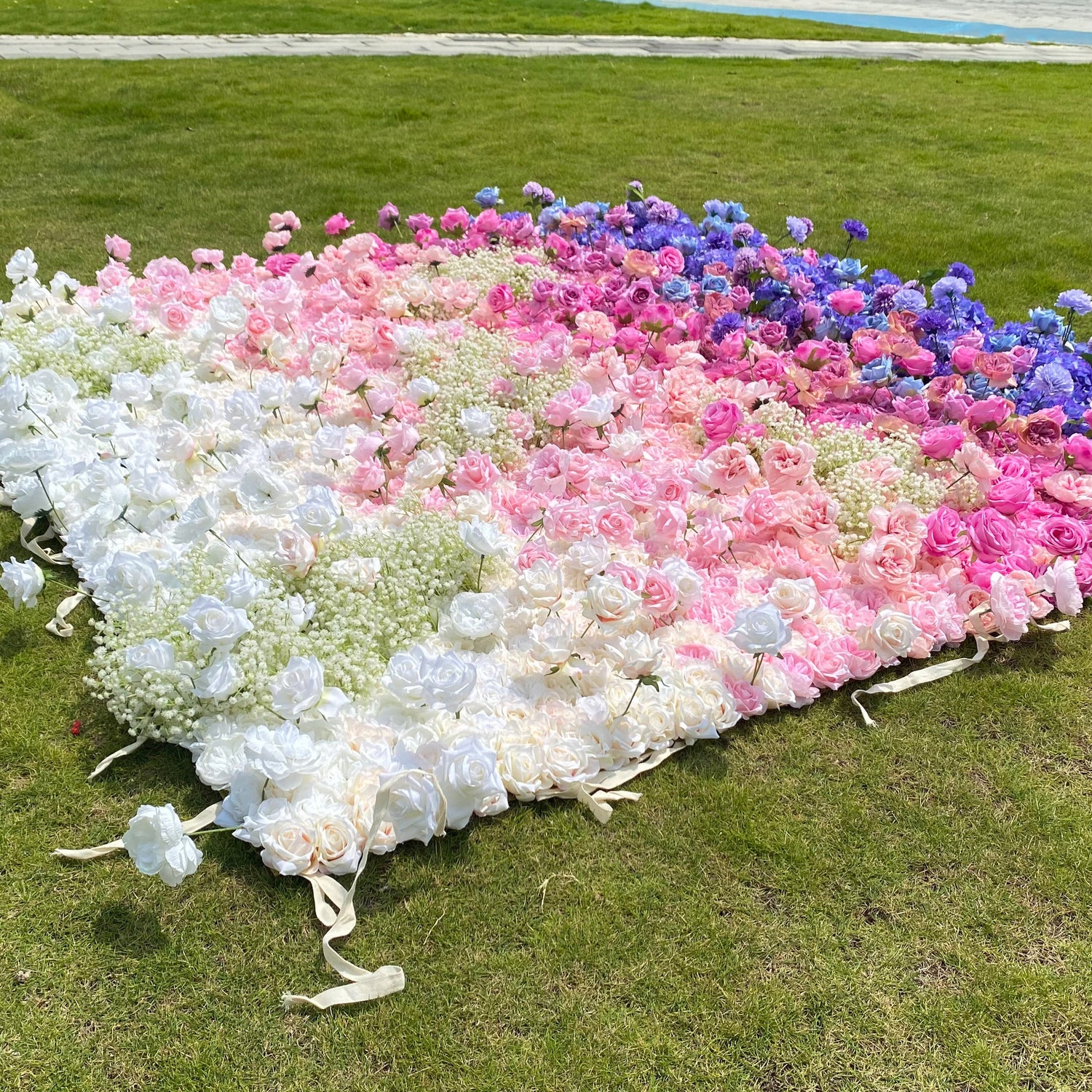 This photo showcases a large floral arrangement laid out on green grass. It features a gradient of white, pink, and blue roses, with baby&
