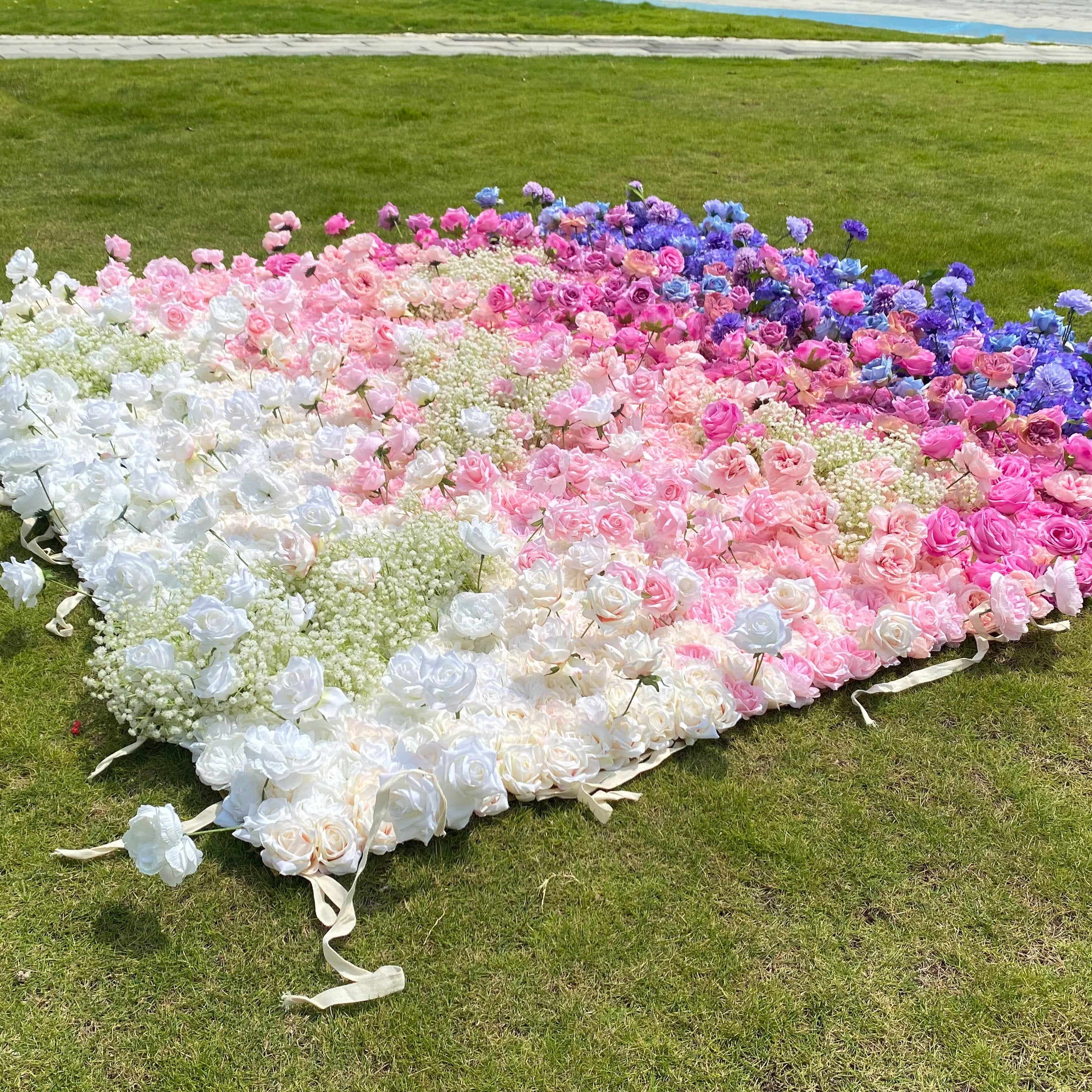 This photo showcases a large floral arrangement laid out on green grass. It features a gradient of white, pink, and blue roses, with baby&