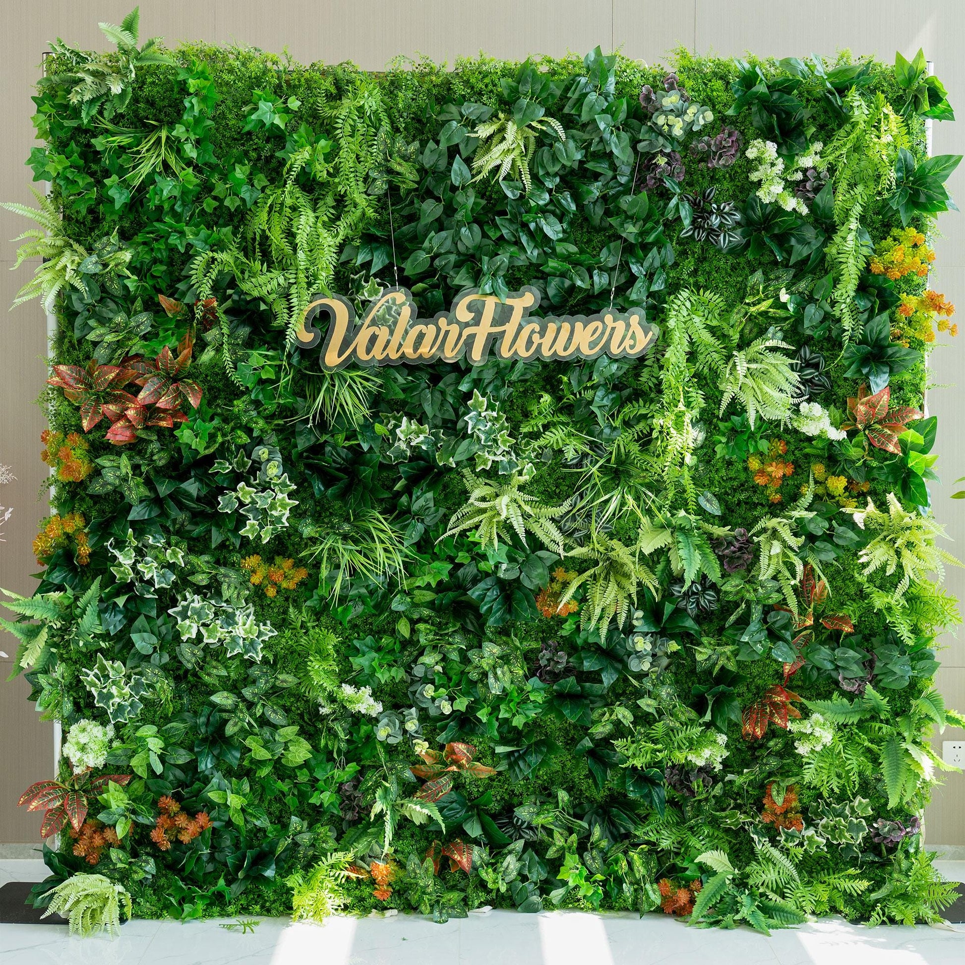 This image showcases a lush, vibrant green wall crafted by ValarFlowers. The wall is densely packed with a variety of artificial plants, featuring different shades of green foliage, including ferns and broad - leafed plants. Splashes of color come from orange, red, and white - tinged plants, adding visual interest. The overall effect is a rich, natural - looking backdrop, ideal for event decoration or as a photo - worthy feature. 