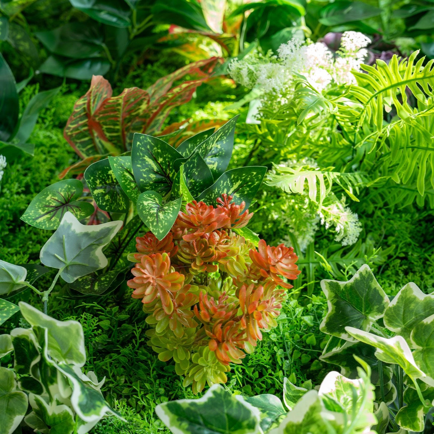This image showcases a vibrant arrangement of artificial plants, likely part of a green wall or backdrop. Prominent are ferns with bright green fronds, adding a lush, natural feel. There are also variegated leaves with green and white patterns, and striking red - and - green coleus - like plants. Additionally, some white, flower - like clusters and ivy - like foliage contribute to the rich, diverse look. 