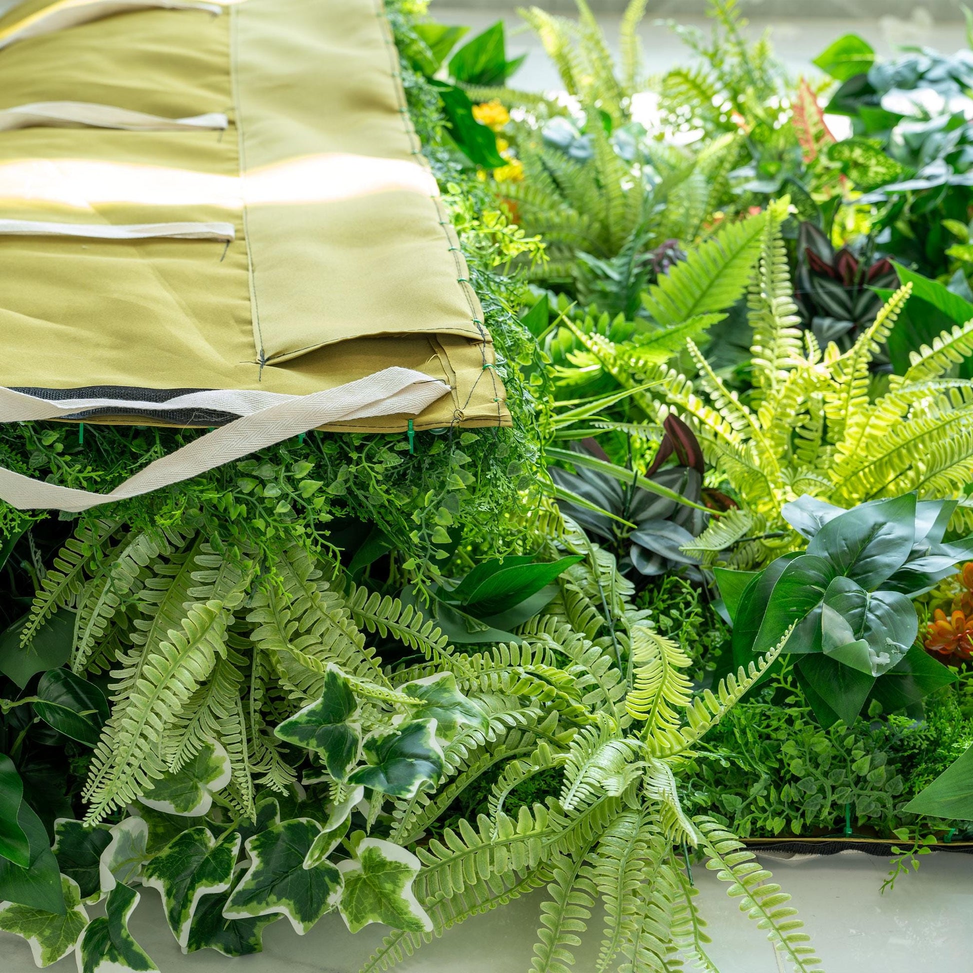 This image shows a close - up view of a lush artificial greenery arrangement. The greenery includes various types of ferns with light and dark green hues, as well as some variegated ivy - like leaves. A mustard - colored fabric with white straps is partially visible, suggesting it might be part of a backdrop or a container for the greenery. The overall scene is vibrant and rich with plant textures. 