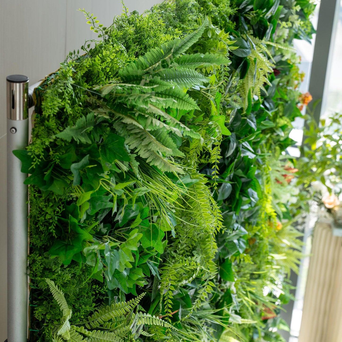 This image showcases a lush, artificial green wall. It features a variety of greenery, including different types of ferns with varying shades of green, from light to dark. The foliage appears dense and vibrant, creating a natural - looking backdrop. A metallic pole is visible on the left side, possibly part of the structure supporting the green wall. The overall scene exudes a fresh and lively atmosphere. 