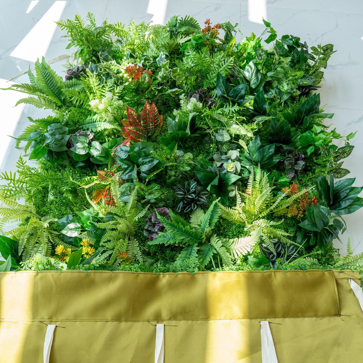 This image features a lush, vibrant green wall densely packed with a variety of artificial plants. The greenery includes different shades of green ferns, broad - leafed plants, and some with variegated patterns. Splashes of color come from red - veined leaves and small orange - hued plants. The bottom of the wall is bordered by a mustard - colored fabric, adding a distinct contrast to the rich greenery above. 