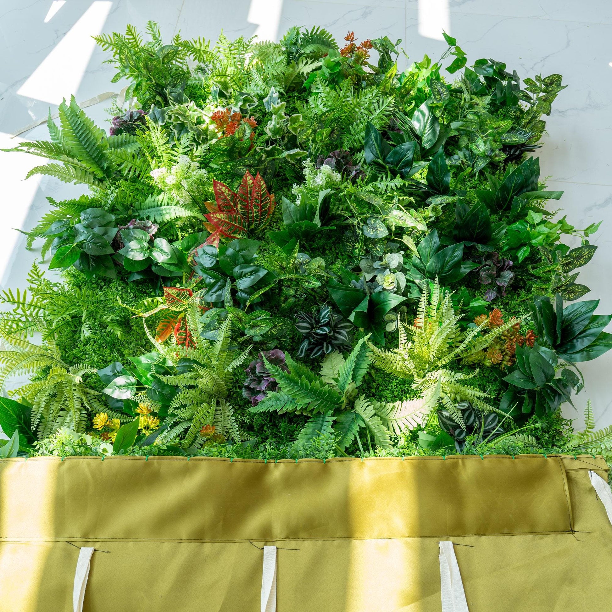 This image features a lush, vibrant green wall densely packed with a variety of artificial plants. The greenery includes different shades of green ferns, broad - leafed plants, and some with variegated patterns. Splashes of color come from red - veined leaves and small orange - hued plants. The bottom of the wall is bordered by a mustard - colored fabric, adding a distinct contrast to the rich greenery above. 