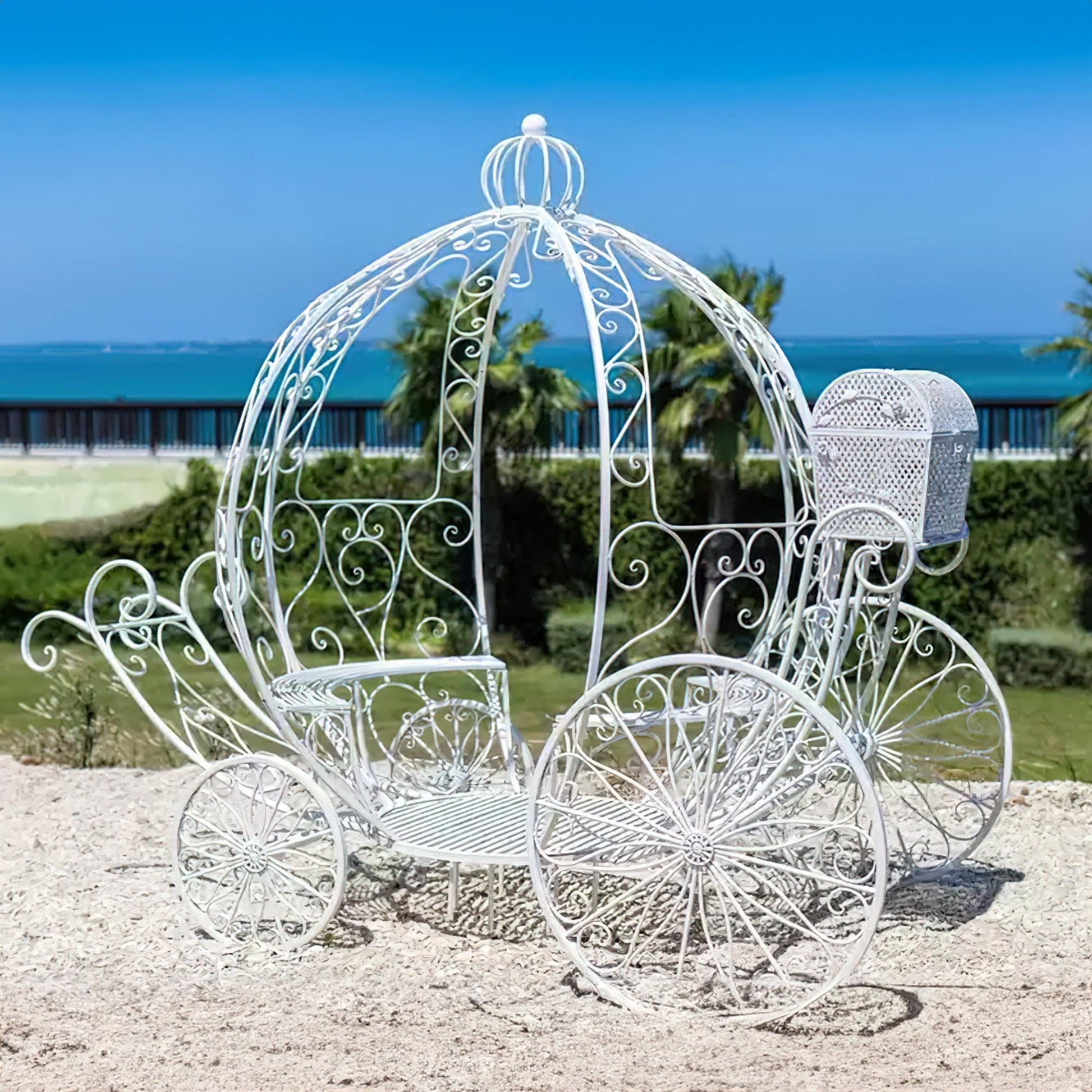 This enchanting white carriage-shaped metal decor by 'ValarFlowers' evokes fairytale charm, featuring intricate scrollwork and a seaside backdrop, perfect for weddings or elegant outdoor events.