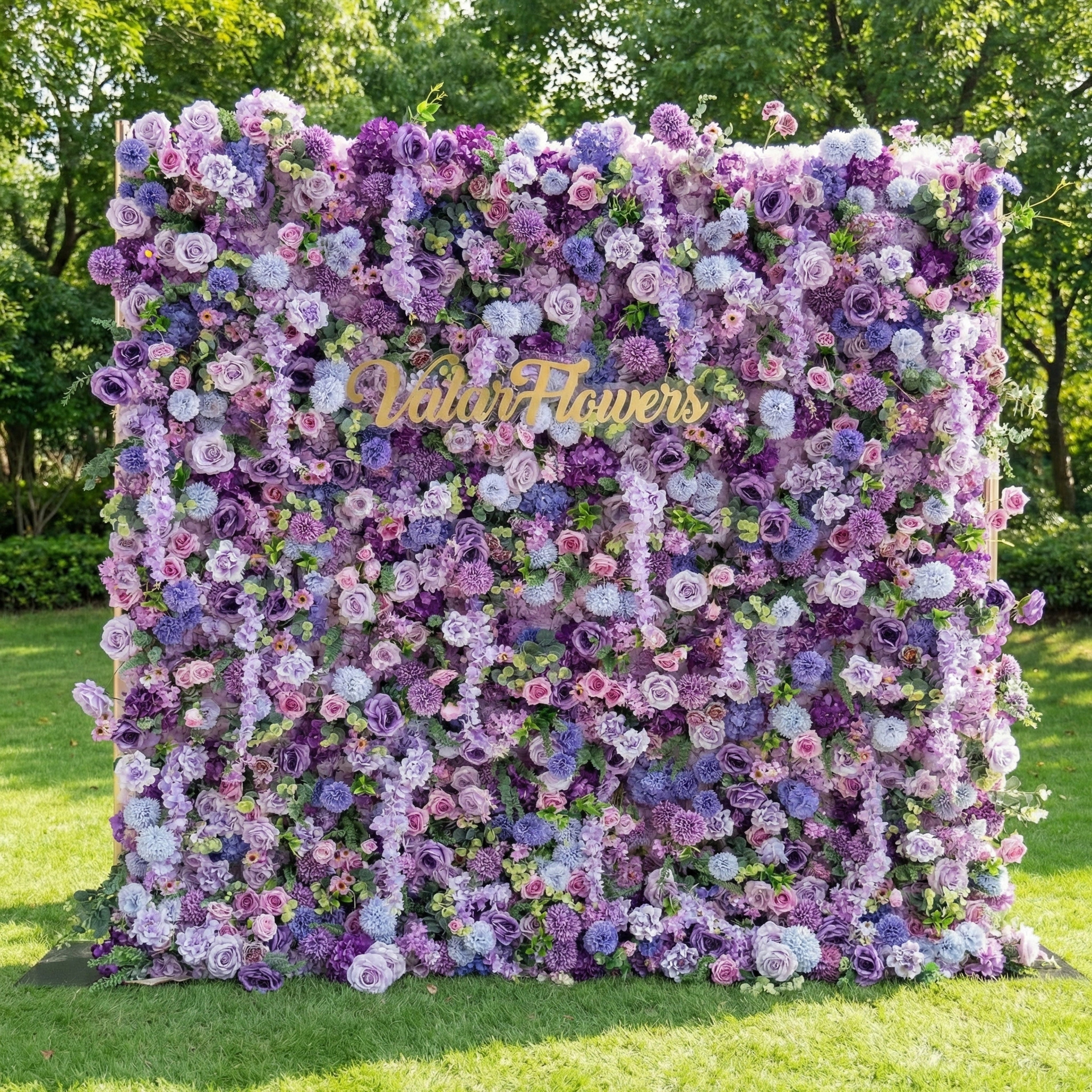 This lush outdoor floral wall from Valar Flowers is a dense tapestry of artificial blooms in dreamy lavender, lilac, and soft blue hues, layered with delicate white and blush-pink roses. Set on a sunlit green lawn framed by leafy trees, its rich, textured arrangement exudes timeless elegance and whimsy, making it a stunning focal point for weddings, garden parties, or photoshoots seeking a soft, romantic aesthetic.