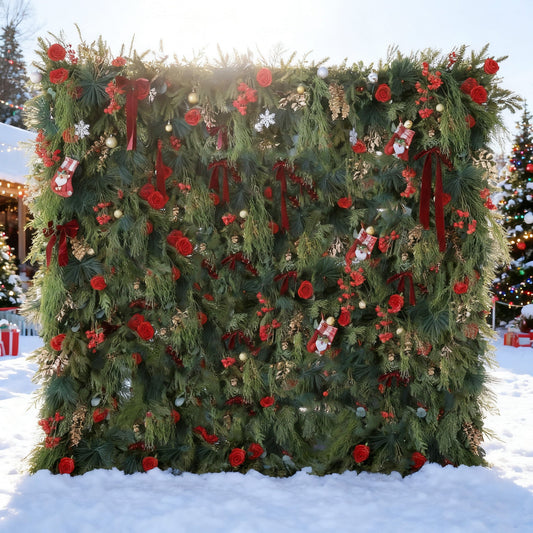 This image features a large, festive Christmas - themed floral wall. Adorned with lush green foliage, vibrant red roses, and Christmas decorations like stockings, snowflakes, and baubles, it's set in a snowy outdoor scene. In the background, lit Christmas trees enhance the joyful, wintry holiday atmosphere.