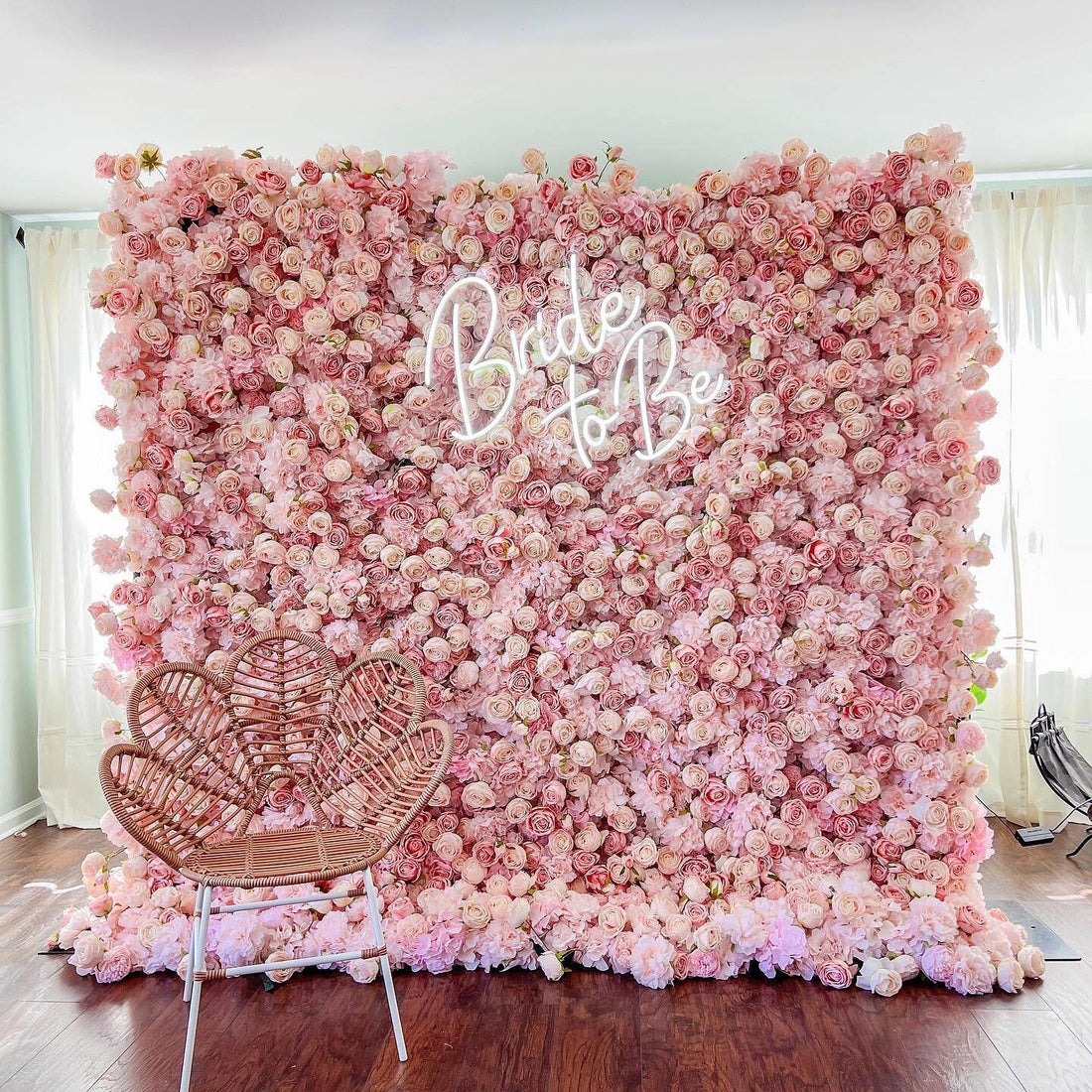 This photo showcases a stunning floral backdrop densely covered in soft pink roses. The word “Bride to Be” is prominently displayed in elegant neon lights, adding a celebratory touch. In the foreground, a heart - shaped wicker chair with a woven design sits on a dark wooden floor, complementing the romantic setting. Softly lit by natural light filtering through sheer curtains, the scene exudes charm and is perfect for bridal events.