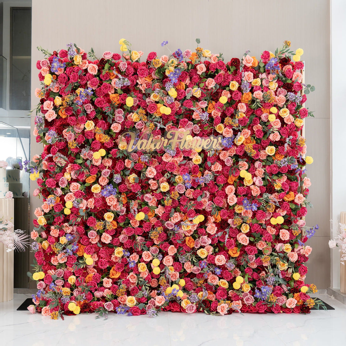 This image showcases a vibrant floral wall densely packed with artificial flowers. Predominant flowers are roses in shades of deep red, pink, and peach. Accents include yellow pom - pom flowers and small purple blooms. The word "ValarFlowers" in cursive script stands out. The style is lush and colorful, ideal for events or photo backdrops. 
